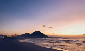 Atardecer en la costa San Sebastián. El cielo se tiñe de tonos rosados y naranjas sobre el mar y la playa, con la silueta del monte Urgull al fondo.