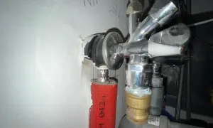Chromglänzendes Eckventil der Wasserleitung mit rotem Schutzschlauch und sichtbaren Leitungen an einer weißen Wand.