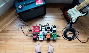 Stojący na drewnianej podłodze pedalboard z efektami gitarowymi, gotowy do gry. W tle wzmacniacz i gitara.