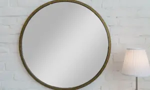 Okrągłe lustro w złotej ramie na białej ceglanej ścianie. Idealne do wieszania lustra bez wiercenia. Obok stoi lampa.
