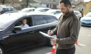 Examinador anotando en una carpeta. Un joven conductor espera en un coche negro. El examen práctico DGT y su precio son importantes para obtener el carnet.