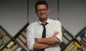 Wojciech Malajkat w białej koszuli i krawacie, z okularami na nosie, stoi na tle regałów z książkami. Jego filmy to klasyka polskiego kina.