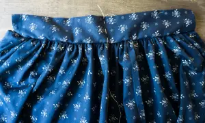 Ein Teil einer dunkelblauen Dirndlschürze mit weißem Blumenmuster wird genäht. Der Bund ist mit Nadeln und Faden fixiert.