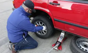 Mężczyzna w niebieskiej kurtce i czapce z daszkiem wykonuje wymianę koła w czerwonym samochodzie, używając podnośnika.