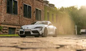 Biała Toyota GR Supra zaparkowana przed ceglanym budynkiem porośniętym bluszczem. Słońce przebija się przez drzewa, tworząc ciepłą, złotą poświatę.