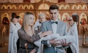 Młodzi rodzice trzymają dziecko do chrztu prawosławnego. W tle widać cerkiew i duchownych.