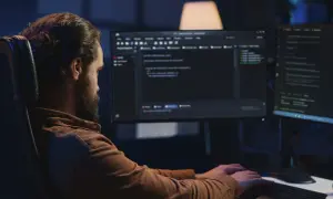 Mężczyzna z brodą, jako java developer, intensywnie pracuje przy dwóch monitorach, pisząc kod w ciemnym pomieszczeniu.