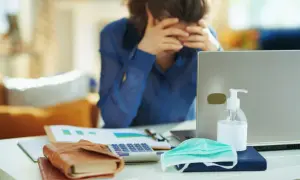 Pracownik przy biurku, zrezygnowany, obok laptop, maseczka, płyn do dezynfekcji, kalkulator. Może to być sytuacja związana z art. 53 kodeksu pracy.