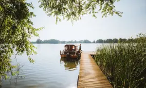 Ein Mann steht auf einem Holzsteg neben einem Hausboot auf einem See. Perfekter Urlaub in Brandenburg.