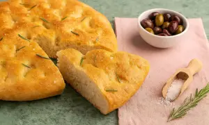 Goldbraune Focaccia, ein Stück davon abgetrennt, mit Rosmarin und grobem Salz bestreut. Daneben eine Schale Oliven und eine Holzschaufel mit Salz. Ein echtes original focaccia rezept.