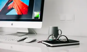 Na biurku iMac, klawiatura, myszka, notatnik z okularami i kubek. Higiena pracy to dbanie o porządek i komfort.