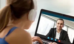Kobieta w słuchawkach pisze na laptopie, prowadząc wideokonferencję z uśmiechniętym mężczyzną. On też ma słuchawki, ale ona pisze ale się nie umawia.