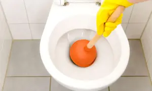 Mano con guanto giallo usa uno stantuffo arancione per liberare un filo d'acqua che scende nel water.
