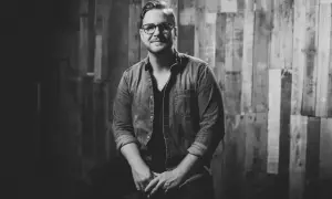 Jordan Reynolds, a talented songwriter, poses confidently in a denim shirt against a rustic wooden backdrop.