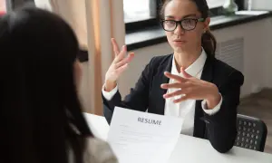 Kobieta w okularach i czarnej marynarce prowadzi rozmowę kwalifikacyjną, gestykulując rękami nad dokumentem "RESUME".