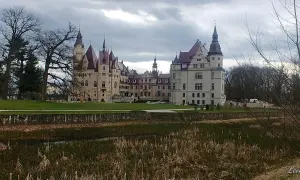 Majestatyczny zamek w Głogówku z wieżami i czerwonymi dachami, otoczony zielonym trawnikiem i stawem z trzcinami.