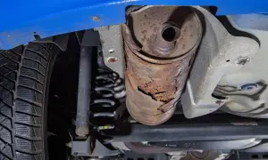 Vue du dessous d'une voiture bleue, montrant une roue, un ressort de suspension et un silencieux rouillé et endommagé.
