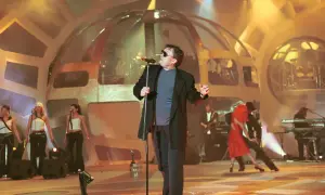Wokalista w okularach śpiewa na scenie, a po nocy przychodzi dzień. W tle tancerze i zespół muzyczny.