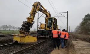 Grupa ludzi w kamizelkach "OPERATOR" obserwuje pracę żółtej koparki przy torach kolejowych. Operator koparki kod zawodu wykonuje swoje obowiązki.