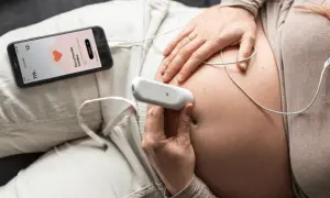 Manos de mujer embarazada usando un doppler fetal para escuchar el latido del bebé. La pantalla del móvil muestra "Escuchando latidos".