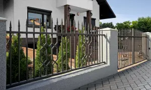 Elegancka brama przesuwna czy dwuskrzydłowa? Metalowe ogrodzenie z ozdobnymi elementami i cyprysami przed domem.