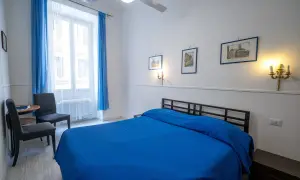 Pokój z niebieską pościelą, idealny na tanie noclegi w Rzymie u Polaków. Okno z niebieskimi zasłonami, dwa krzesła i stolik.