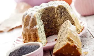 Pyszna babka makowa, oprószona cukrem pudrem, z widocznym ziarnem maku. Obok miseczka z makiem i kawałek ciasta.