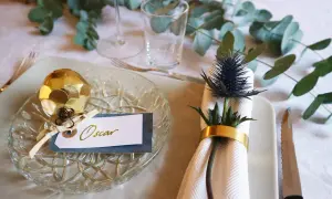 Elegant nyårsdukning med gulddekorationer och en namnskylt för Oscar. En gyllene kotte pryder tallriken, medan en blå tistel med guldfärgad servettring skapar en vacker detalj.