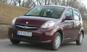 Ciemnoczerwony Subaru Justy jedzie po drodze, zalesione wzgórze w tle.