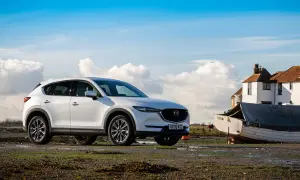 Vit Mazda CX-5 parkerad vid en båt på en lerig strand, med hus och moln i bakgrunden.