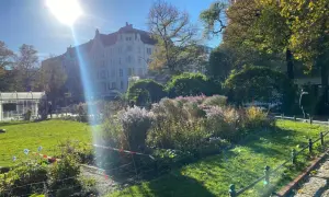 Grüner Park mit Blumenbeeten, Bäumen und einem Altbau unter strahlender Sonne.