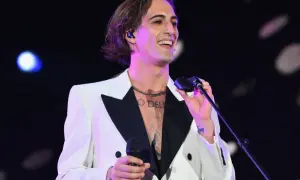 Damiano David w białej marynarce z czarnym kołnierzem śpiewa na koncercie, trzymając mikrofon.