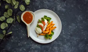 Talerz z batatami, brokułami, mozzarellą i pomidorem, obok łyżka z sosem.