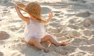 Liten bebis på stranden med solskyddsfaktor, leker med en stor stråhatt.