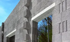 Ściana z bloczków betonowych z widocznym betonowym nadprożem nad otworem okiennym. W tle zbrojenie i drzewa.