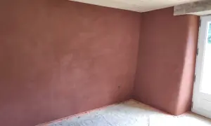 Un mur d'angle avec un enduit chaux sable intérieur d'une belle teinte ocre, sous un plafond en bois clair. Le sol est protégé par une bâche.
