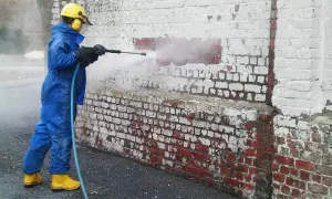 Personne en combinaison bleue et bottes jaunes utilise un nettoyeur haute pression pour enlever peinture sur brique.