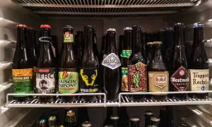 Scaffale pieno di bottiglie di birra, alcune con etichette colorate come "Zinnebir" e "Saison Dupont". Una birra aperta in frigo, pronta per essere gustata.