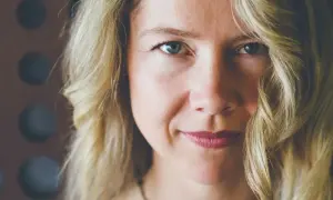 Zbliżenie na twarz kobiety z blond włosami. Delikatny uśmiech, lekko opadające policzki, niebieskie oczy.