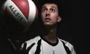 Młody siatkarz w białej koszulce z napisem ALLSIX przygotowuje się do challenge w siatkówce, unosząc piłkę nad dłonią.