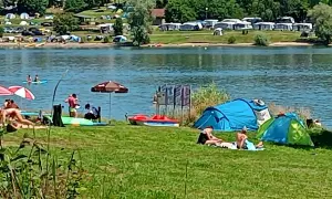 Letnie popołudnie nad jednym z malowniczych **jezior w Czechach**. Ludzie odpoczywają na trawie, opalają się, a w wodzie pływają kajaki. W tle widać pole namiotowe.