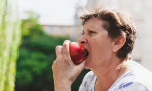 Starsza kobieta gryzie soczyste jabłko, zastanawiając się, czy jabłka tuczą. Zdjęcie z bliska, na zewnątrz.