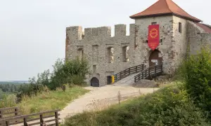 Kamienne mury zamku w Rabsztynie, z drewnianym mostkiem prowadzącym do bramy. Czerwony baner zaprasza na wydarzenie.