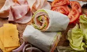 Dwa zawinięte w papier wrapy z szynką, serem, pomidorami i sałatą. Obok plastry szynki, sera, pomidorów i cebuli. Idealne na szybki posiłek, choć warto sprawdzić wrap mcdonald kcal.