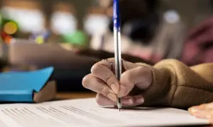 Dłoń pisząca długopisem po papierze, w tle rozmyte sylwetki osób.