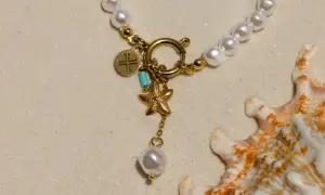 Un delicado llavero de oro con charms de estrella de mar y perla, sobre arena y junto a una concha marina.