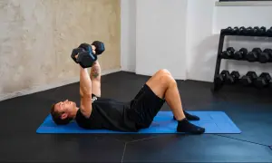Uomo esegue esercizi pettorali con manubri su tappetino blu. Allenamento in palestra con rack di pesi sullo sfondo.
