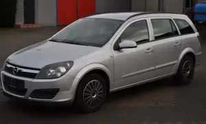 Srebrny Opel Astra H 1.6 benzyna, kombi, zaparkowany na kostce brukowej przed budynkiem z czerwonymi drzwiami.