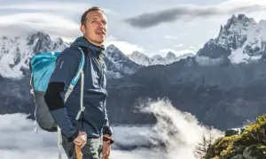 Randonneur souriant, sac à dos bleu et bâtons de marche, prêt à affronter le poids de la montagne. Paysage alpin spectaculaire.