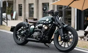 Ciemnozielony motocykl typu bobber z brązowym siedzeniem, zaparkowany na ulicy.